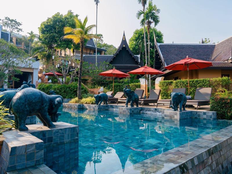Ein Pool mit Steinfiguren von Tieren und roten Sonnenschirmen in einer grünen Gartenanlage.
