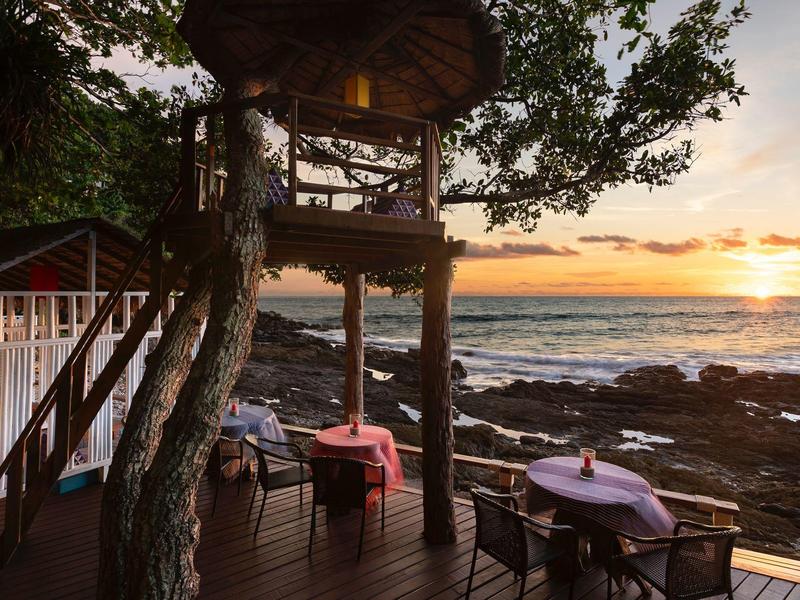 Terrasse mit Holzmöbeln und Baum, Blick auf Meer und Sonnenuntergang am felsigen Ufer.