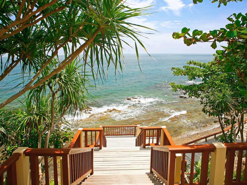 Holztreppe führt durch grüne Pflanzen zum Meer mit blauem Himmel und leicht bewölktem Horizont.