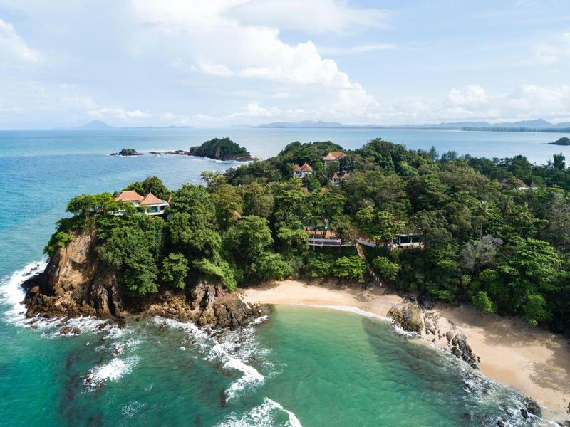 Grüne, bewaldete Insel mit Stränden und Felsen, umgeben von blau-grünem Meer unter bewölktem Himmel.