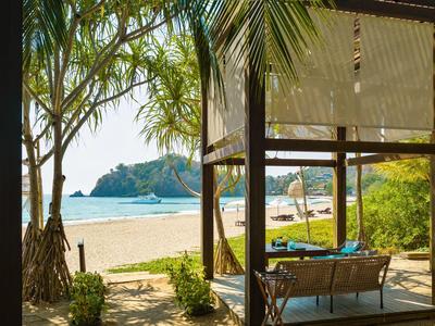 Blick von einer überdachten Lounge am Strand mit Palmen, Sand und klarem Meerwasser.