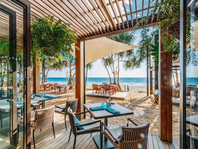 Offener Restaurantbereich mit Holzterrasse, Stühlen und Tischen, Blick auf Strand und Meer
