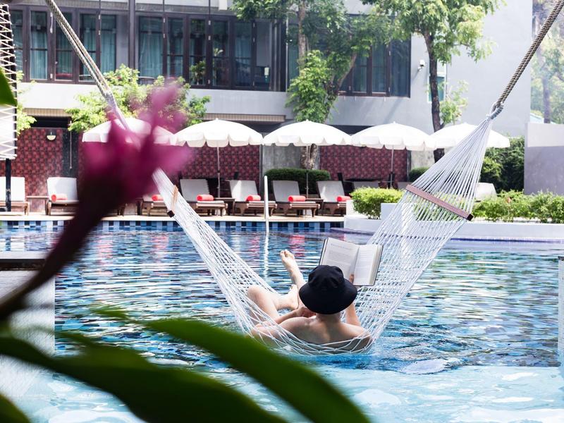 Person entspannt in einer Hängematte über einem Hotelpool mit Liegestühlen und Sonnenschirmen im Hintergrund