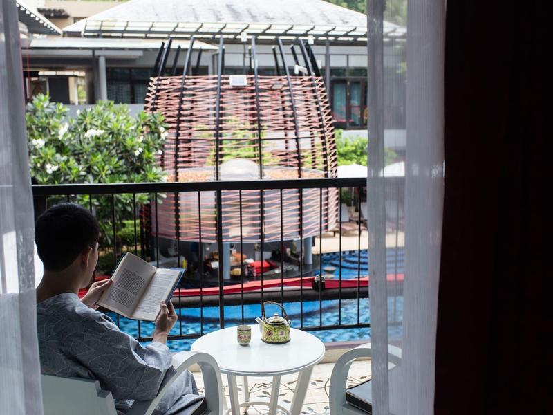 Person sitzt auf Balkon mit Buch, kleiner Tisch mit Tasse, im Hintergrund Pool und Garten mit Pflanzen.