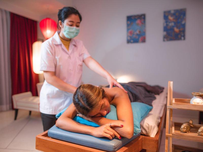 Frau erhält Rückenmassage auf Massagetisch, Therapeutin trägt Maske, entspannte Atmosphäre mit warmem Licht.