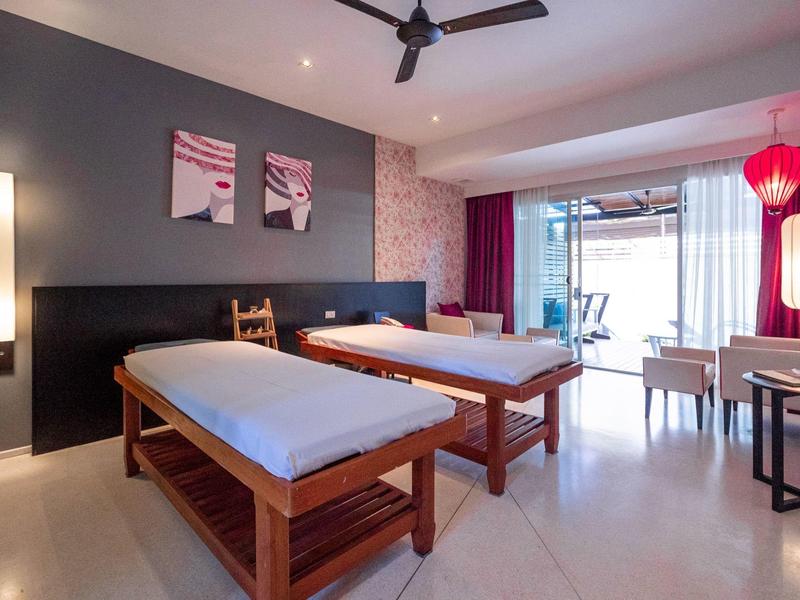 Helles Behandlungszimmer mit zwei Massageliegen, dunkler Wand und großen Fenstern mit roter Deko.