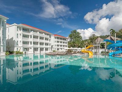 Großer Hotelpool mit Wasserrutschen vor weißem Hotelgebäude unter blauem Himmel.