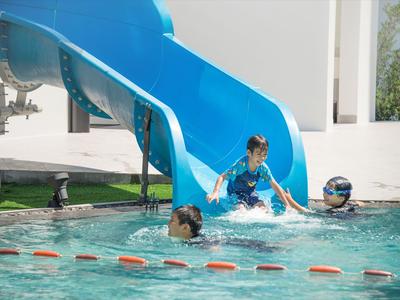 Kinder spielen im Schwimmbecken neben einer blauen Wasserrutsche an einem sonnigen Tag.
