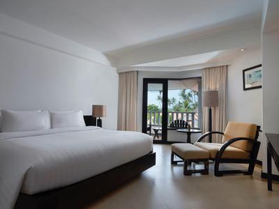 Helles Hotelzimmer mit großem Bett, Ledersessel, Balkon und Blick auf Palmen und Meer.