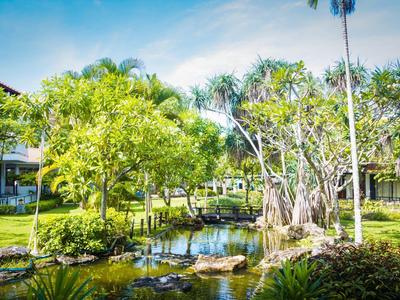 Garten mit Teich, Bäumen, Palmen und grünen Wiesen vor hellen Gebäuden unter blauem Himmel.