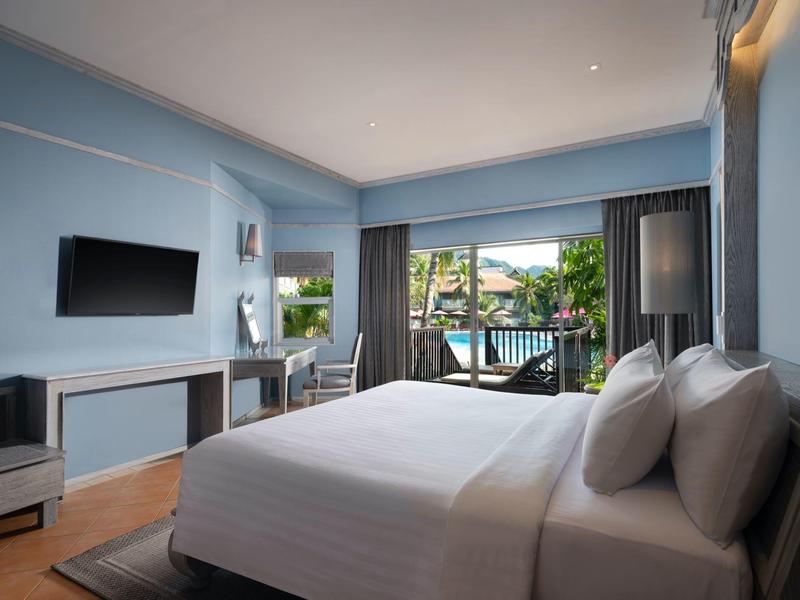 Modernes Hotelzimmer mit großem Bett, Balkon, TV, Schreibtisch und Blick auf Pool und Palmen.