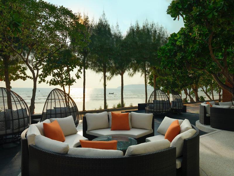 Runde weiße Gartenlounge mit orangen Kissen auf einer Terrasse, umgeben von Bäumen am Wasser.