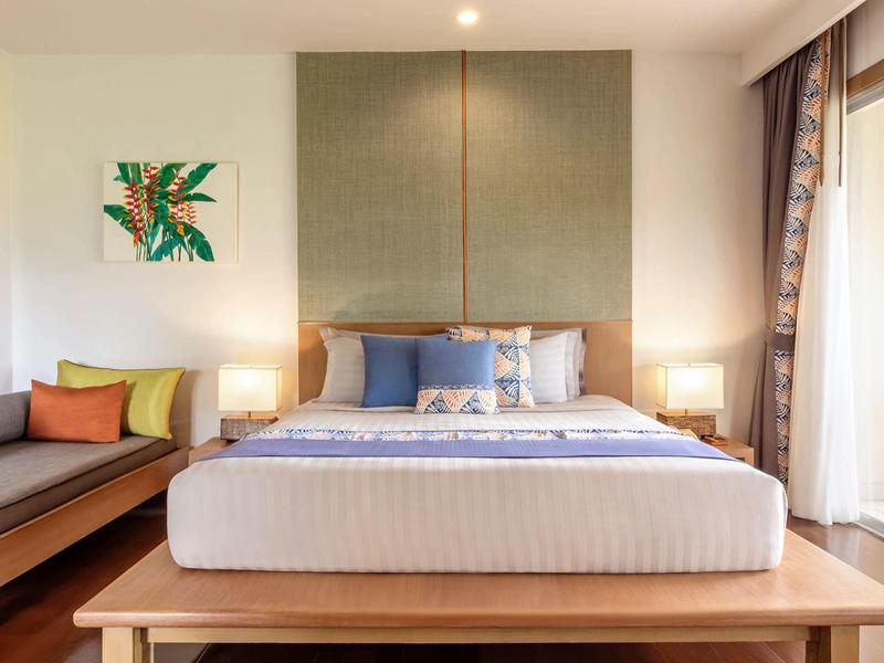 Modernes Schlafzimmer mit großem Bett, Holzbank, Sofa, bunten Kissen und einem abstrakten Wandbild.