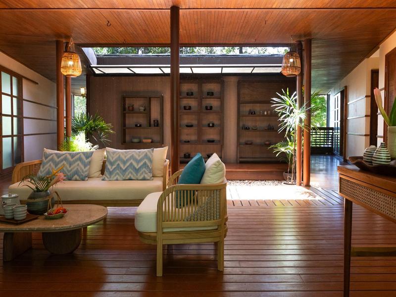 Helles Wohnzimmer mit Holzboden, Rattansofa, Teetisch und großen Pflanzen, offen zum Außenbereich.