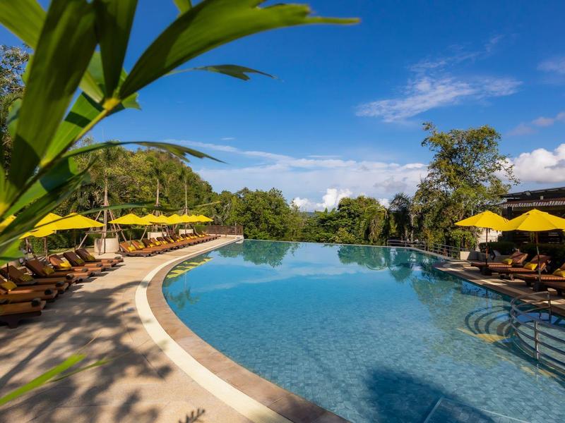 Großer, gebogener Pool mit Liegestühlen, gelben Sonnenschirmen und grüner Umgebung bei blauem Himmel.