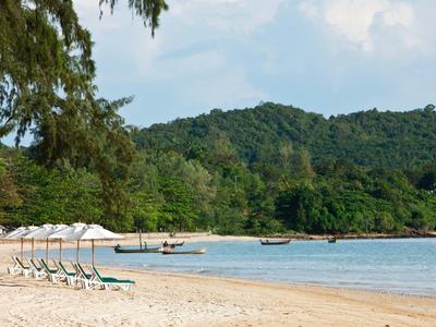 Leerstehender Sandstrand mit weißen Sonnenschirmen, grün bewaldeten Hügeln und blauem Himmel.