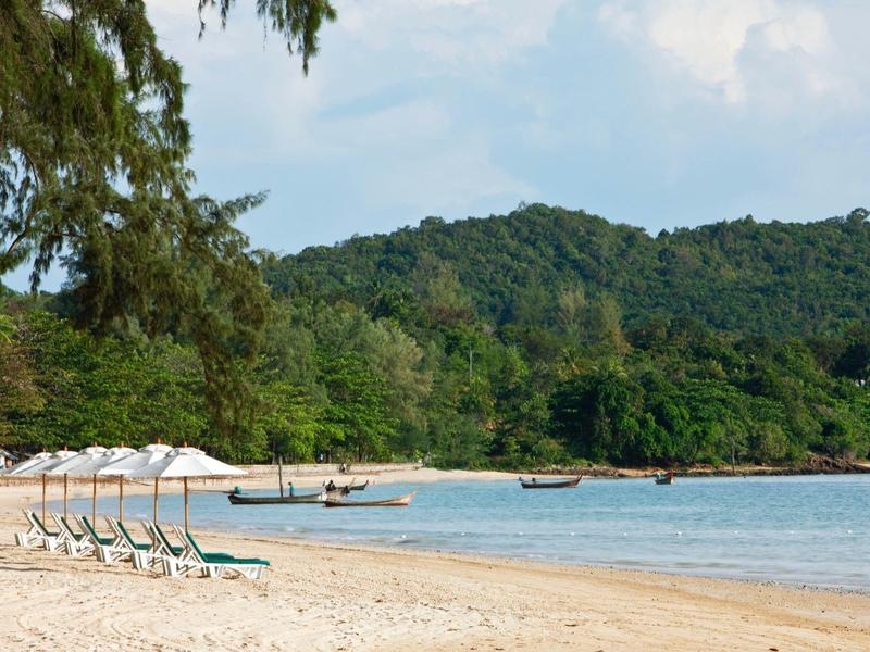 Leerstehender Sandstrand mit weißen Sonnenschirmen, grün bewaldeten Hügeln und blauem Himmel.