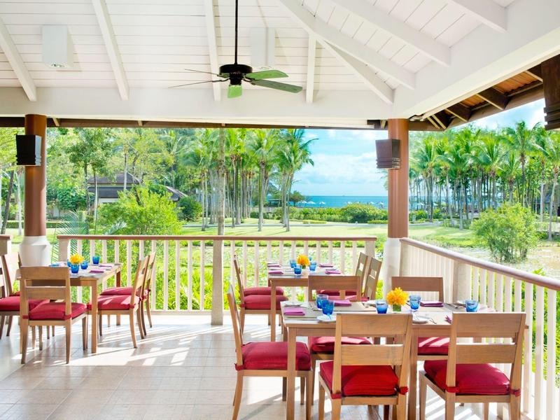 Offener Essbereich mit Holztischen, roten Stühlen und Blick auf grüne Bäume und Meer.