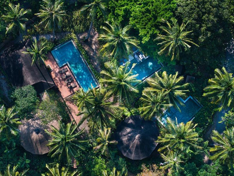 Vogelperspektive eines tropischen Resorts mit Pool, Palmen und runden Strohdächern im dichten Grün.