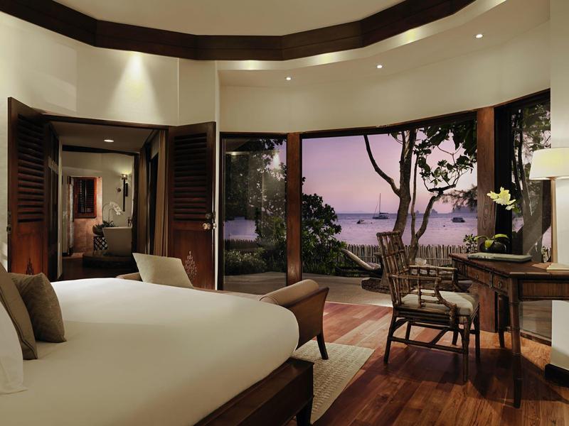 Ein gemütliches Schlafzimmer mit großem Bett, Holzboden und Blick auf Terrasse und Meer bei Sonnenuntergang.