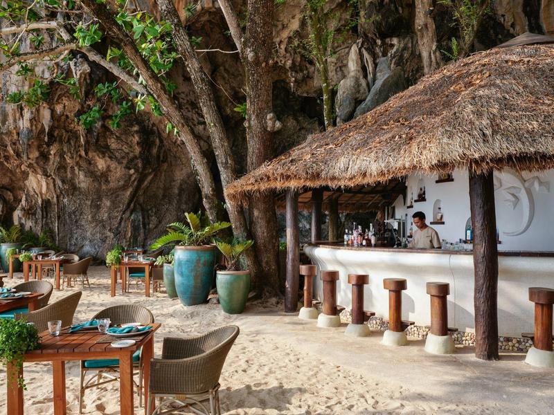 Strandbar mit Strohdach, Holzstühlen, Tischen und Pflanzen vor bewaldetem Felsenstrand.
