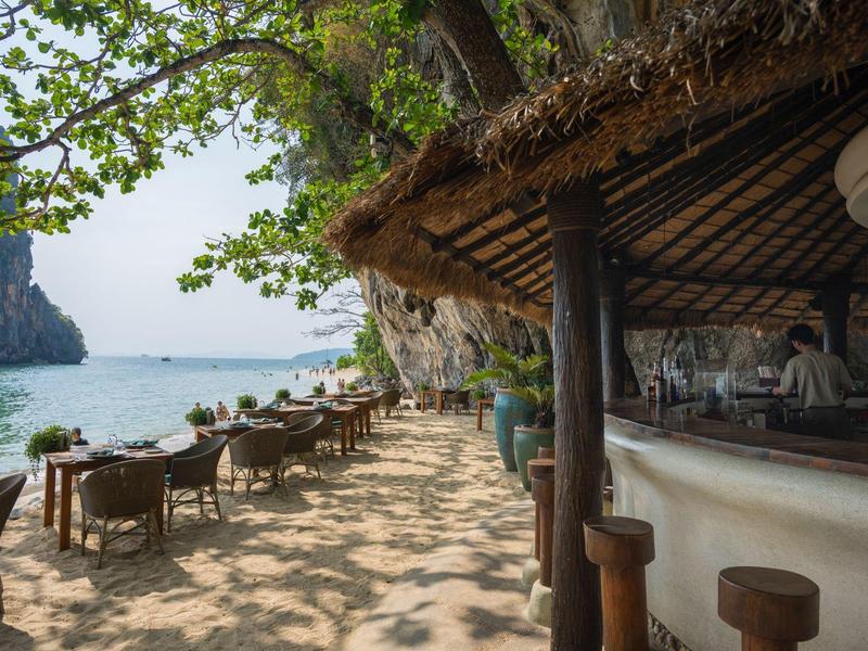 Strandbar mit Strohüberdachung, Sandboden und Tischen unter Bäumen neben klarem Meerwasser.