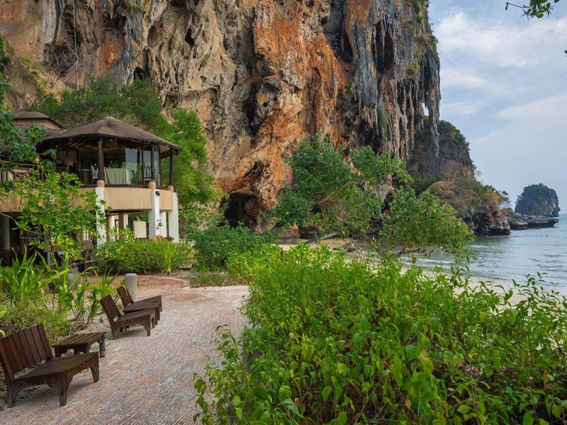 Strand mit Holzbänken, grünem Buschwerk und einem Haus unter Felsen neben dem Meer