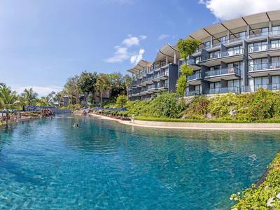 Großer Pool im Freien, umgeben von Liegestühlen, Palmen und mehrstöckigem Hotelgebäude bei blauem Himmel.