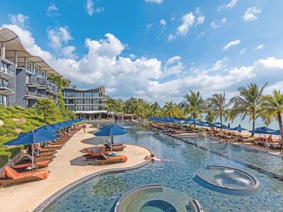 Großer Hotelpool mit Liegestühlen, Sonnenschirmen und Palmen unter blauem Himmel am Meer.