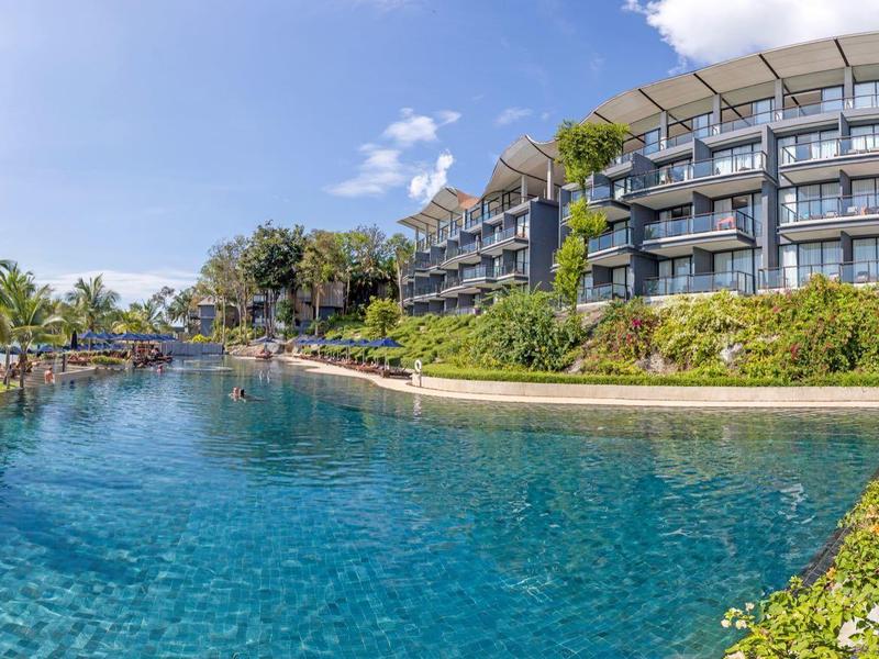 Großer Pool im Freien, umgeben von Liegestühlen, Palmen und mehrstöckigem Hotelgebäude bei blauem Himmel.