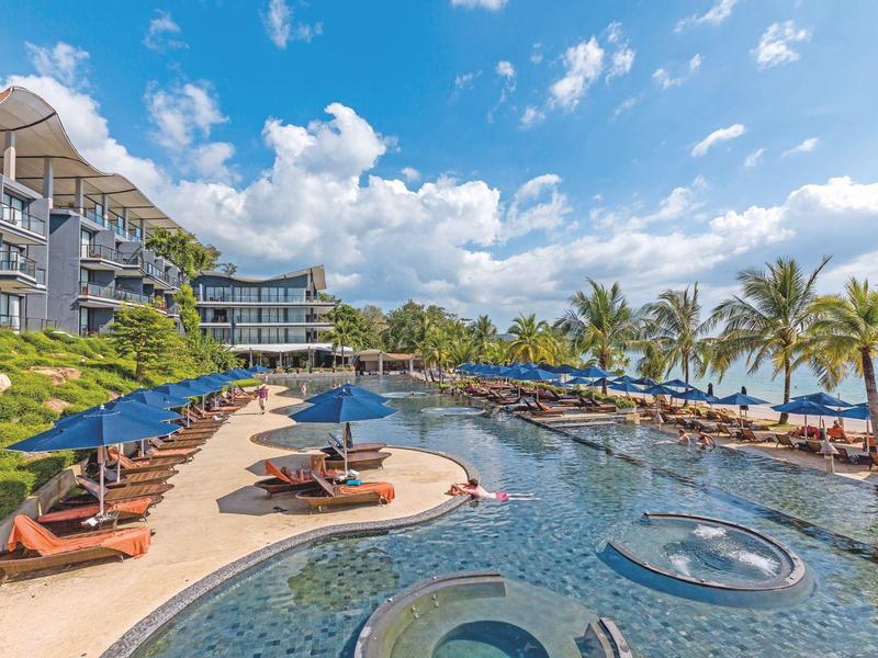 Großer Hotelpool mit Liegestühlen, Sonnenschirmen und Palmen unter blauem Himmel am Meer.