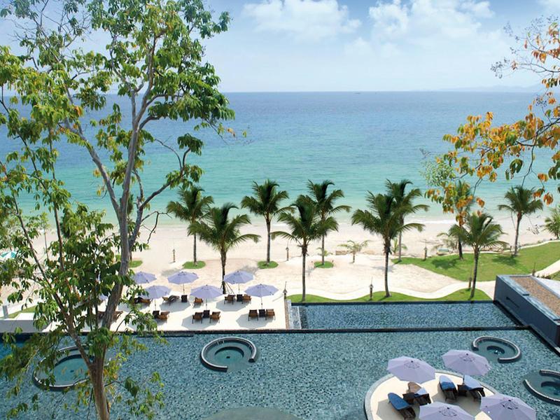 Blick vom Pool mit Palmen, blauem Meer, Sandstrand und Sonnenschirmen unter blauem Himmel.