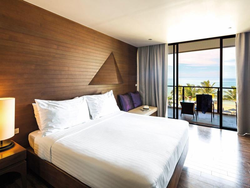 Modernes Schlafzimmer mit großem Bett, Holzwand, Tischlampe und Blick auf Balkon und Meer.