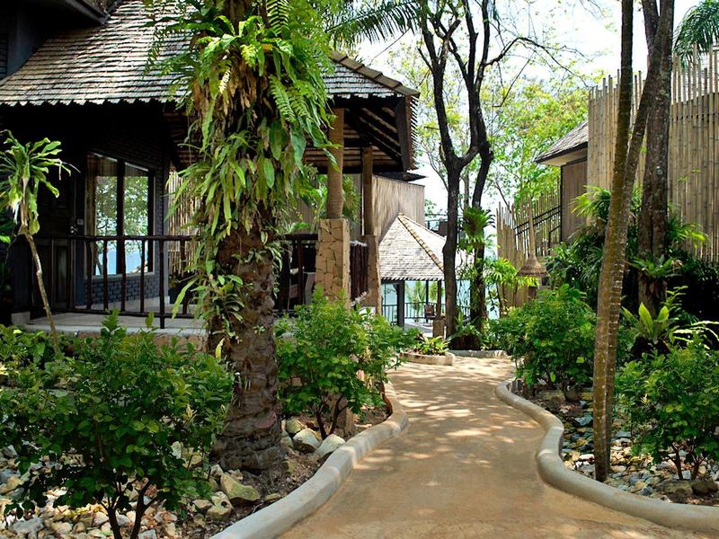 Weg führt durch grünen Garten zu hölzernen Hütten mit tropischen Pflanzen und Bäumen.