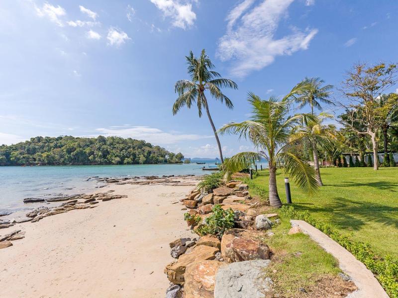 Strand mit Palmen, grünem Gras und einer kleinen Insel im Hintergrund unter blauem Himmel.