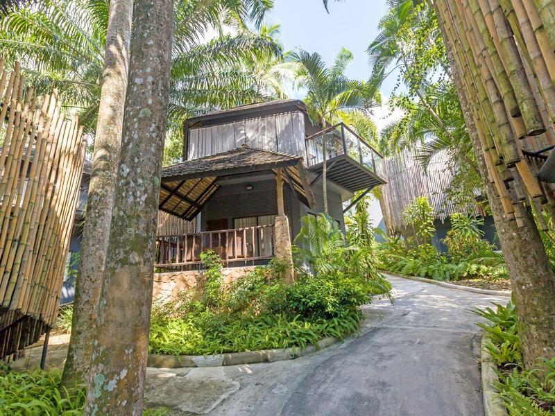 Pfad führt zu einem erhöhten Bungalow aus Holz, umgeben von grüner Vegetation und Palmen.