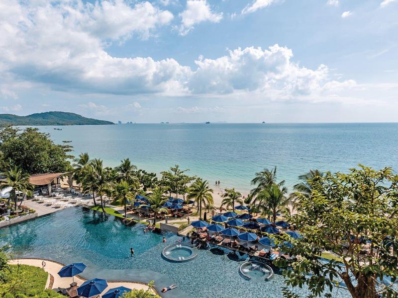 Hotelpool mit blauen Sonnenschirmen und Blick auf den Strand und das Meer unter bewölktem Himmel