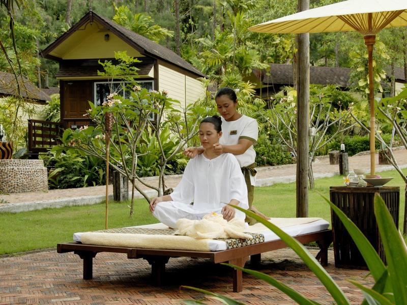 Frau bekommt im Garten eine Nackenmassage auf einer Liege unter Sonnenschirm, umgeben von Pflanzen.