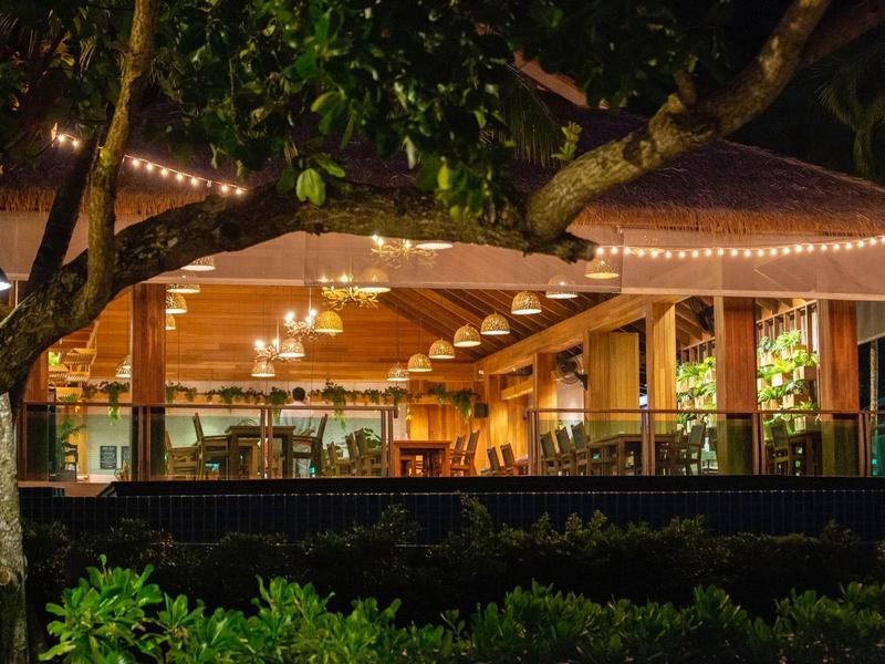 Gemütliches, beleuchtetes Restaurant mit Holzdecke und großem Fenster in tropischer Nacht.