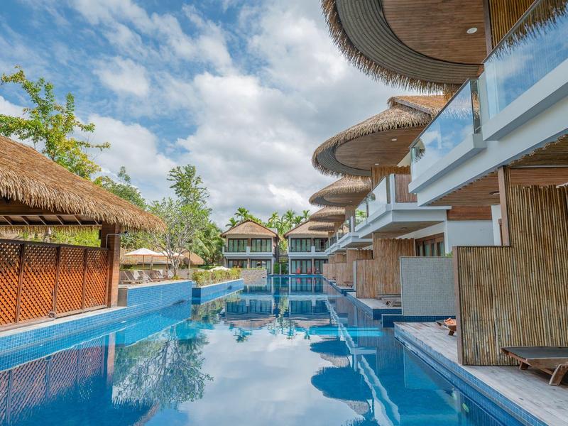 Moderner Pool mit Wasserspielen vor tropischen Bungalows und Sonnenschirmen.