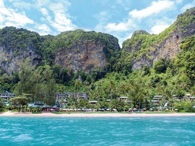 Klares türkisfarbenes Meer vor grüner Küste mit Strand und steilen Felsen unter blauem Himmel.