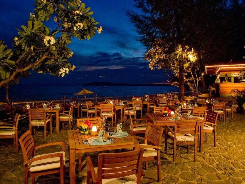 Abendterrasse mit Holztischen, Stühlen, Kerzenlicht und Bäumen bei klarem Nachthimmel am Meer.