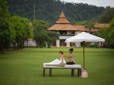 Eine Person erhält eine Rückenmassage auf einer Liege im Garten mit Bäumen und asiatischem Gebäude.