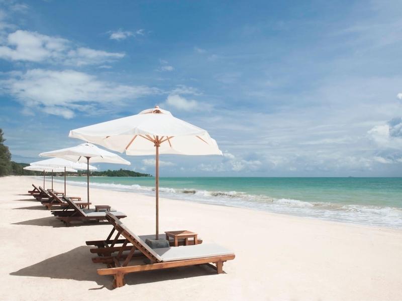 Strand mit weißen Sand, Holzliegen unter weißen Sonnenschirmen und blauem Himmel mit Wolken.