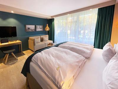Helles Hotelzimmer mit großem Bett, Sofa, Schreibtisch und großen Fenstern mit Gardinen.