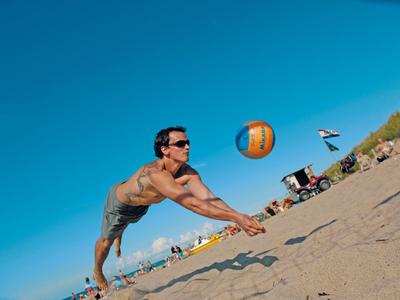A man dives for a volleyball on a sandy beach under a clear blue sky.