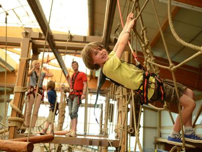 Children climbing on an indoor adventure playground with ropes and wooden elements.