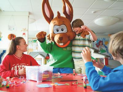 Children and a mascot sit at a red table in a bright room with decorations.