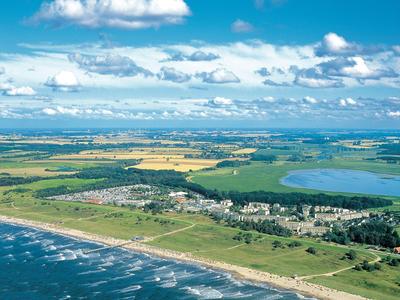 Küste mit Sandstrand, grünen Feldern, Dorf und See unter blauem Himmel mit Wolken.