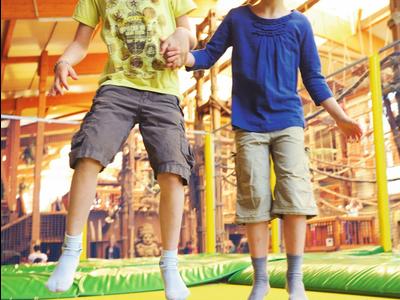 Zwei Kinder springen lachend auf einem grünen und gelben Trampolin in einer Halle.